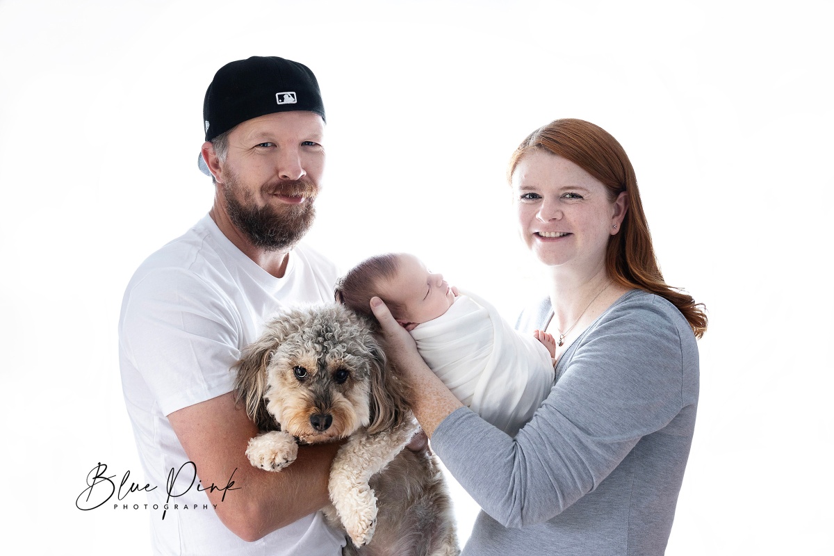 Heartwarming high-key image with a newborn baby cradled in the arms of a loving mother, while the proud father holds their dog, all against a pristine white background. Parents gaze into the camera, capturing a moment of pure joy and family connection.